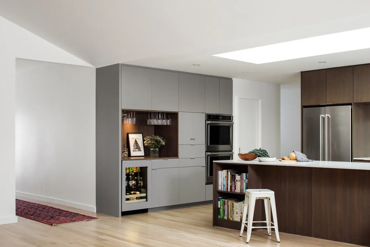 Kitchen with gray cabinets, dark wood island with bookshelves, and double wall ovens under skylight lighting.
