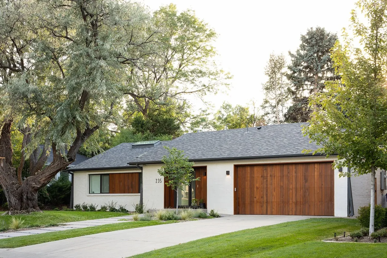 The Krameria Residence features a wood panel garage door and entrance on a white stucco exterior with a shingle roof and concrete stepping stones on the driveway.