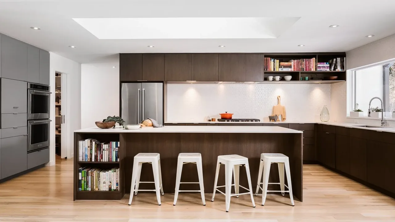 Kitchen of Krameria Residence features a dark wood island with built-in shelving, white stools, and integrated appliances.