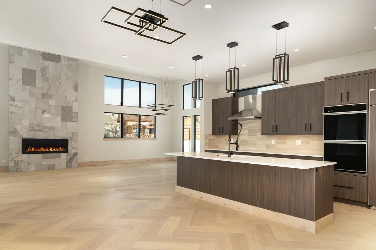 Modern open kitchen with island overlooks spacious living area with stone fireplace and floor-to-ceiling windows.
