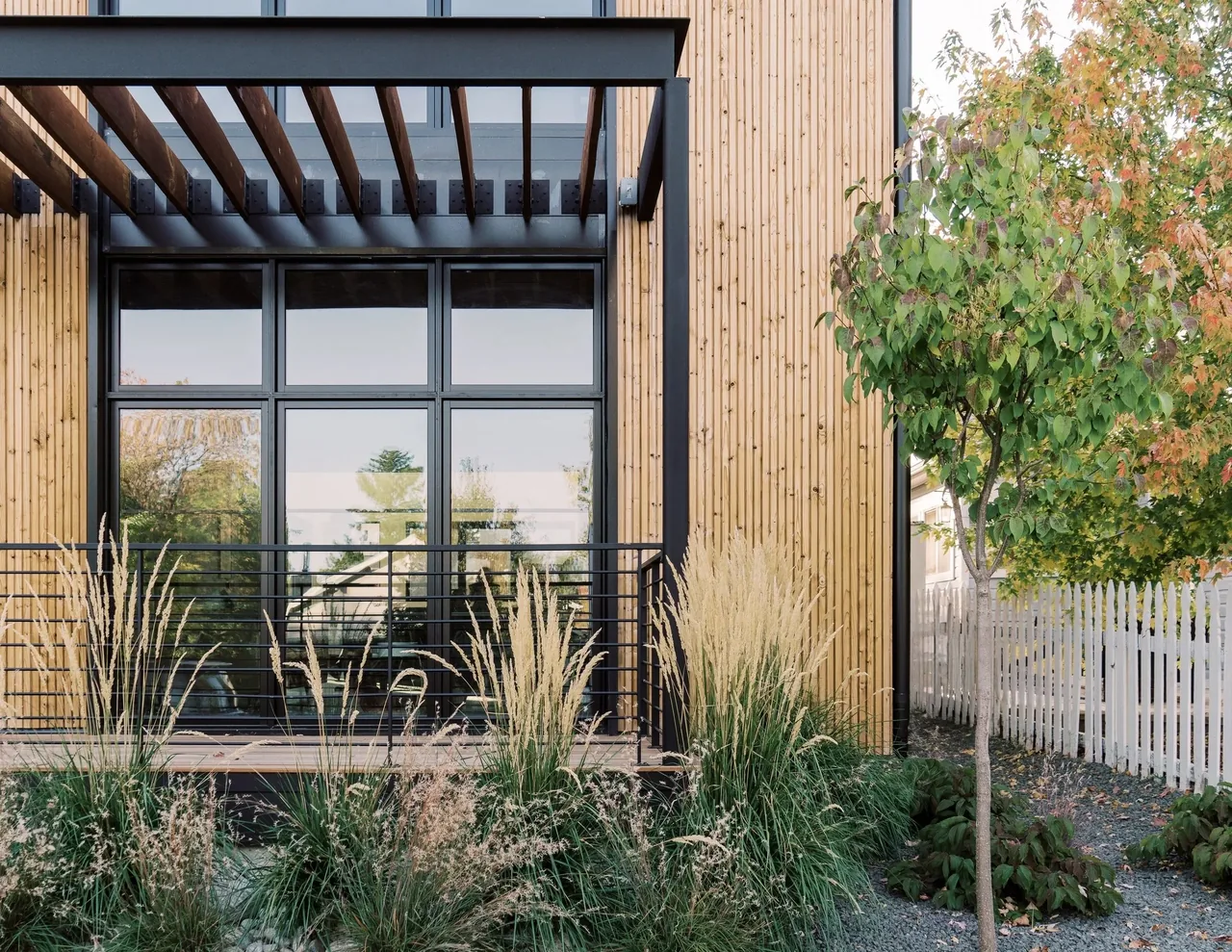 Modern house exterior featuring vertical wood siding, large picture windows, and a black metal pergola over a porch with wire railing.