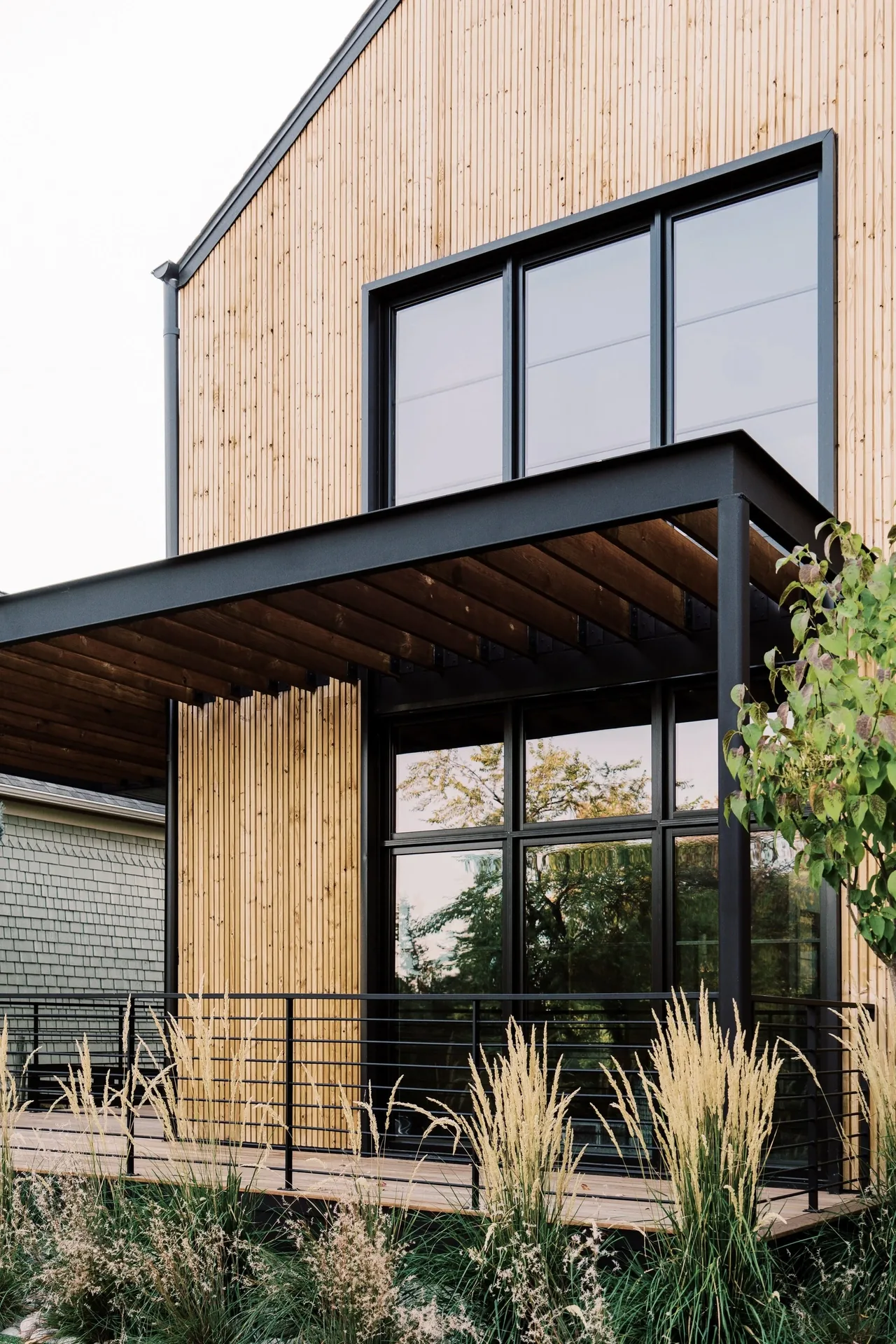 LOUISVILLE NET-ZERO RESIDENCE features vertical wood siding, a black metal pergola, and large picture windows with black frames.