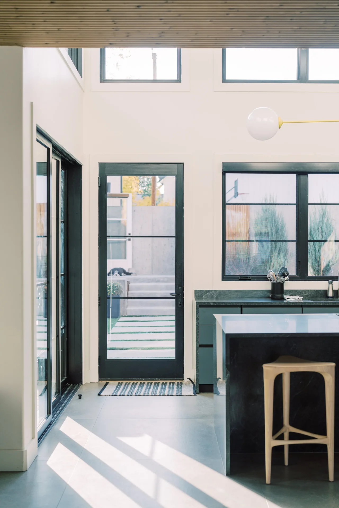 Modern kitchen interior with black framed glass door and windows, kitchen island and natural wood stool in Louisville Net-Zero Residence.