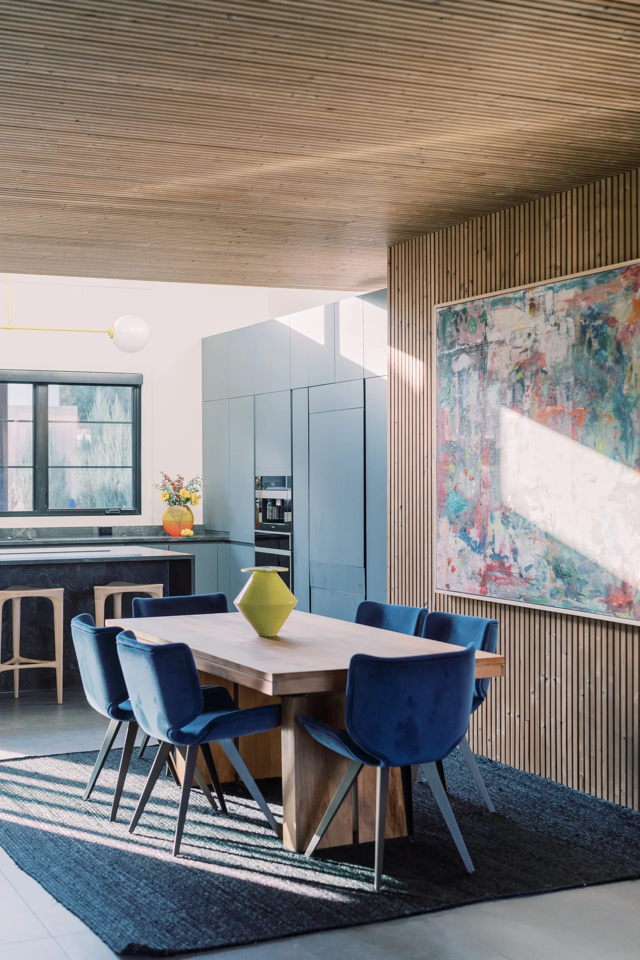 Dining area with blue chairs, wooden table, and adjacent kitchen with gray cabinetry illuminated by natural light.