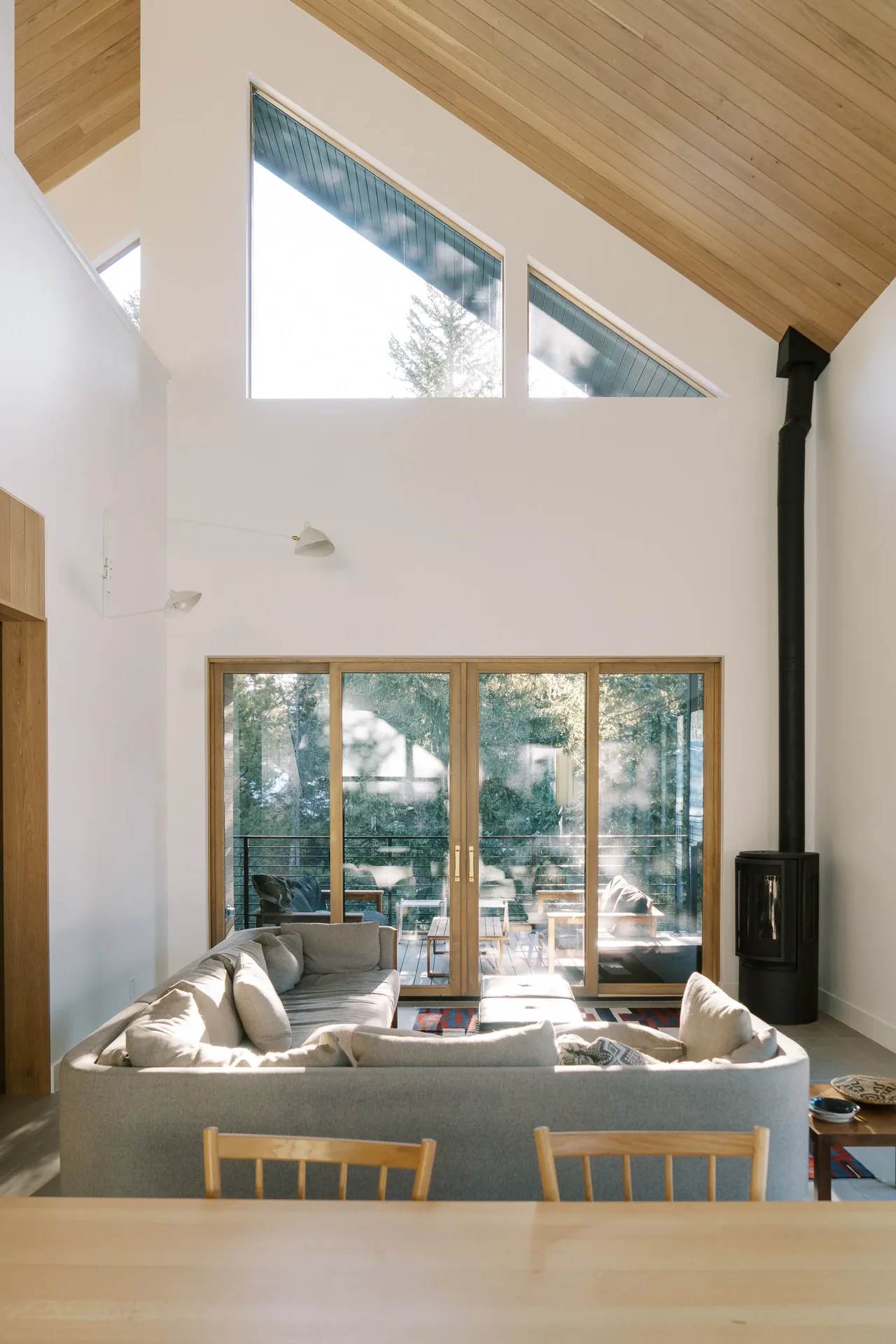 Living area with large triangular windows and sliding glass doors opening to a balcony overlooking forest.
