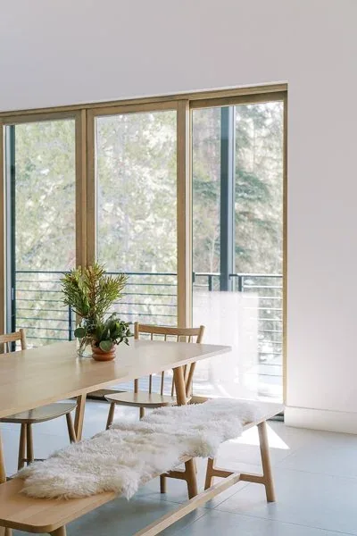 Dining space features natural wood furniture and floor-to-ceiling glazing overlooking an outdoor balcony with metal railing.