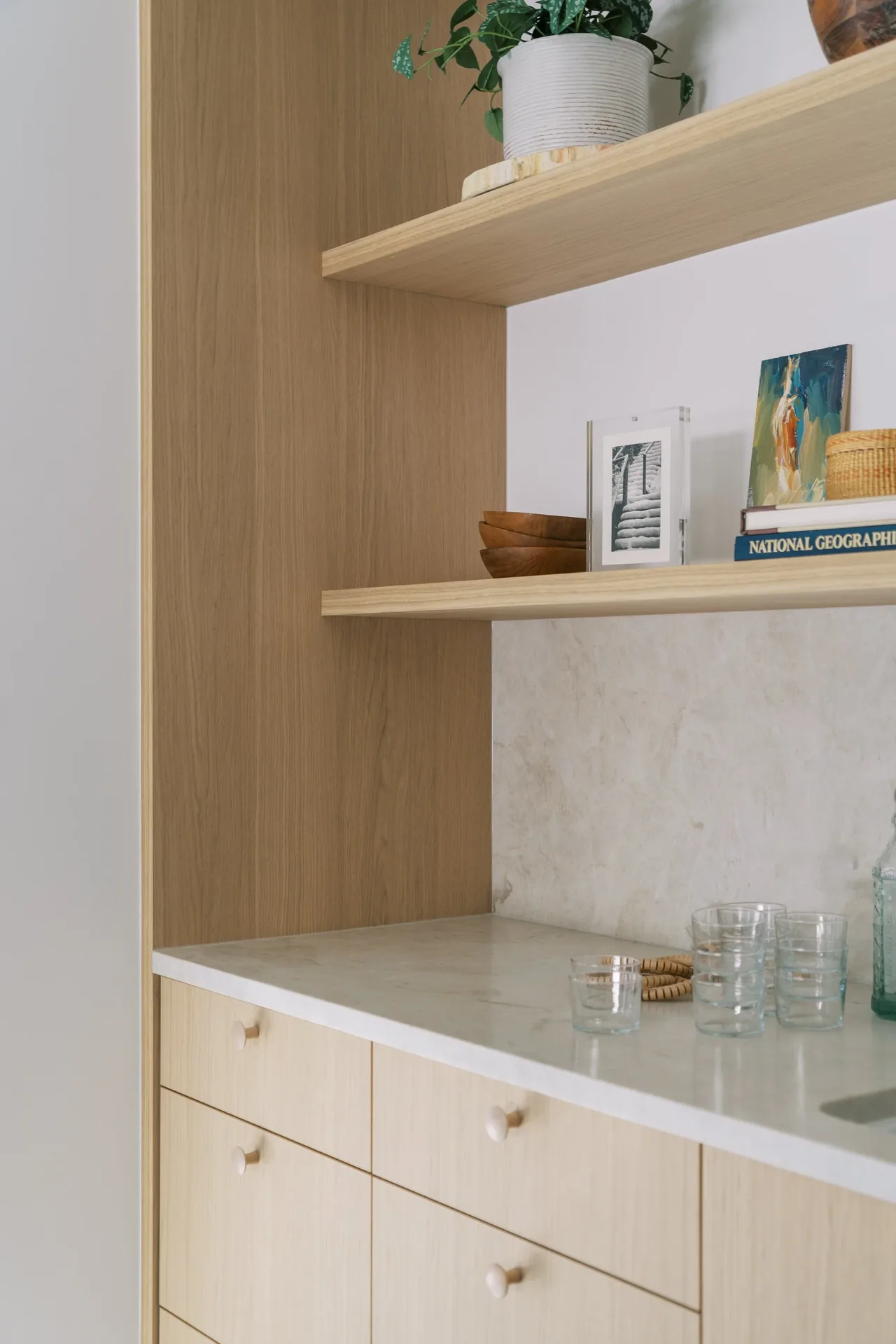 Kitchen cabinetry with a white countertop and light wood finish features floating shelves with decorative items.