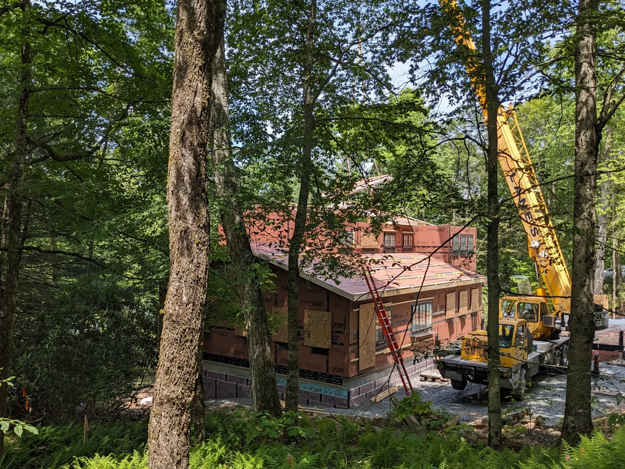 Lake Cottage under construction showing exterior sheathing and roof framing amidst dense forest.