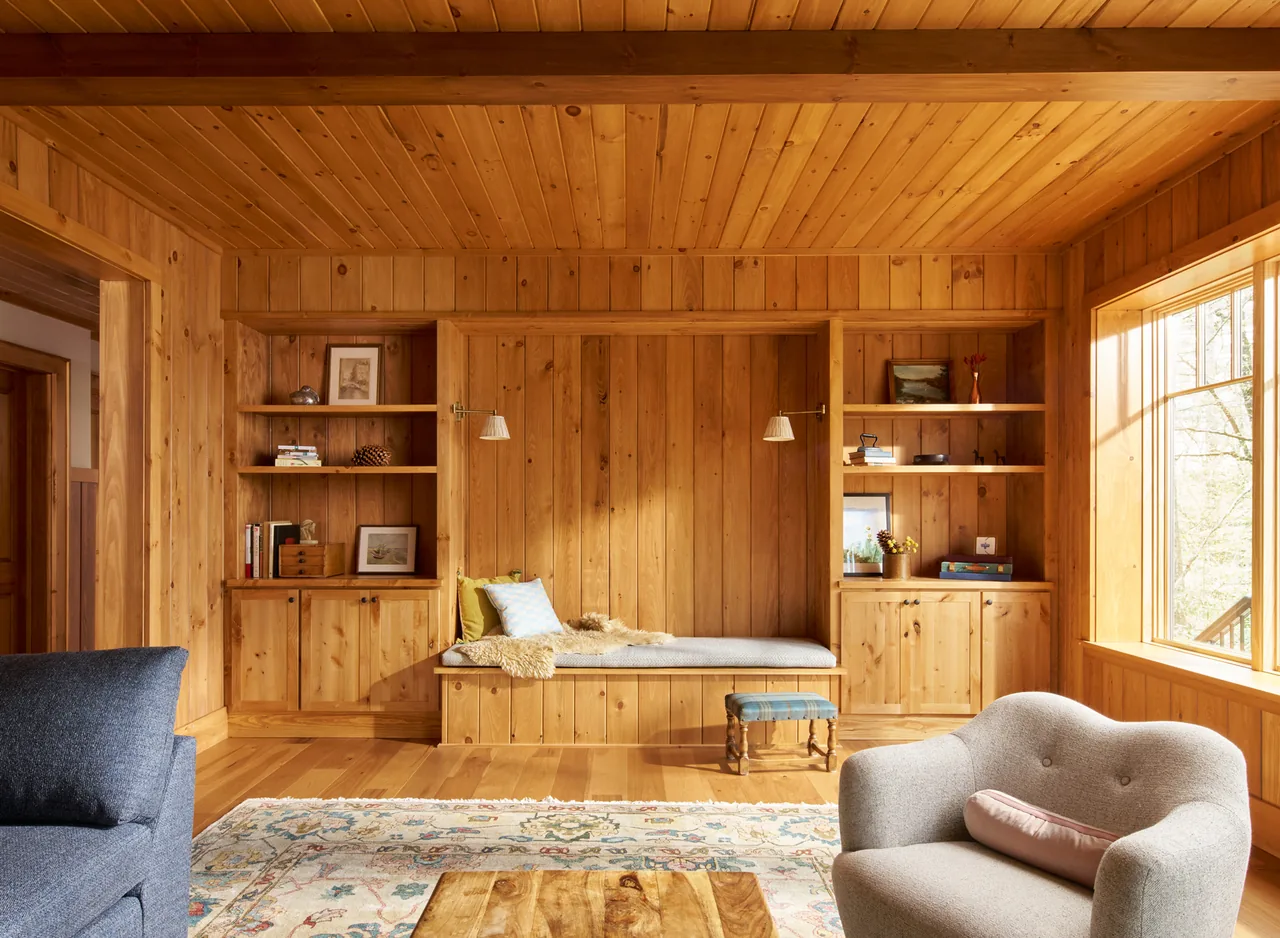 Living room interior featuring wood paneling, built-in shelving and bench, and large window letting in natural light.