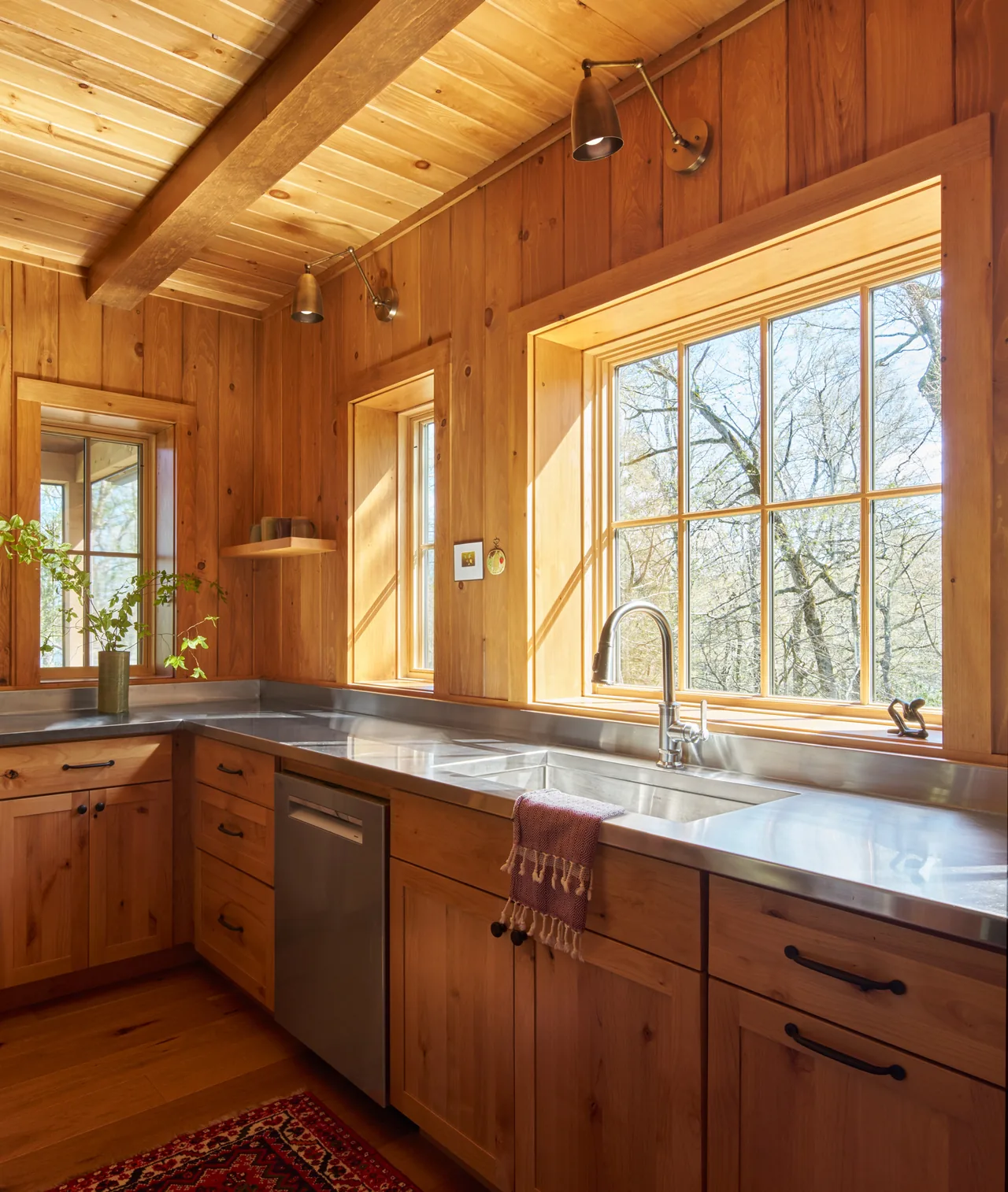 Lake Cottage kitchen features wood panels and stainless steel countertops with a view of bare trees outside.