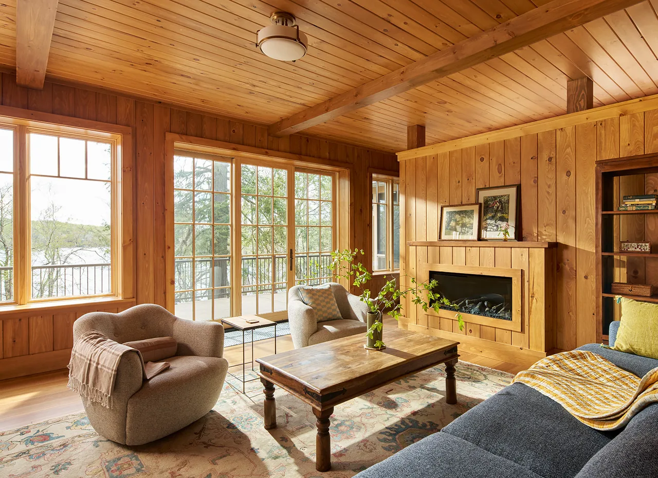 Lake Cottage living room features natural wood paneling, a built-in fireplace, and large windows with lake views.