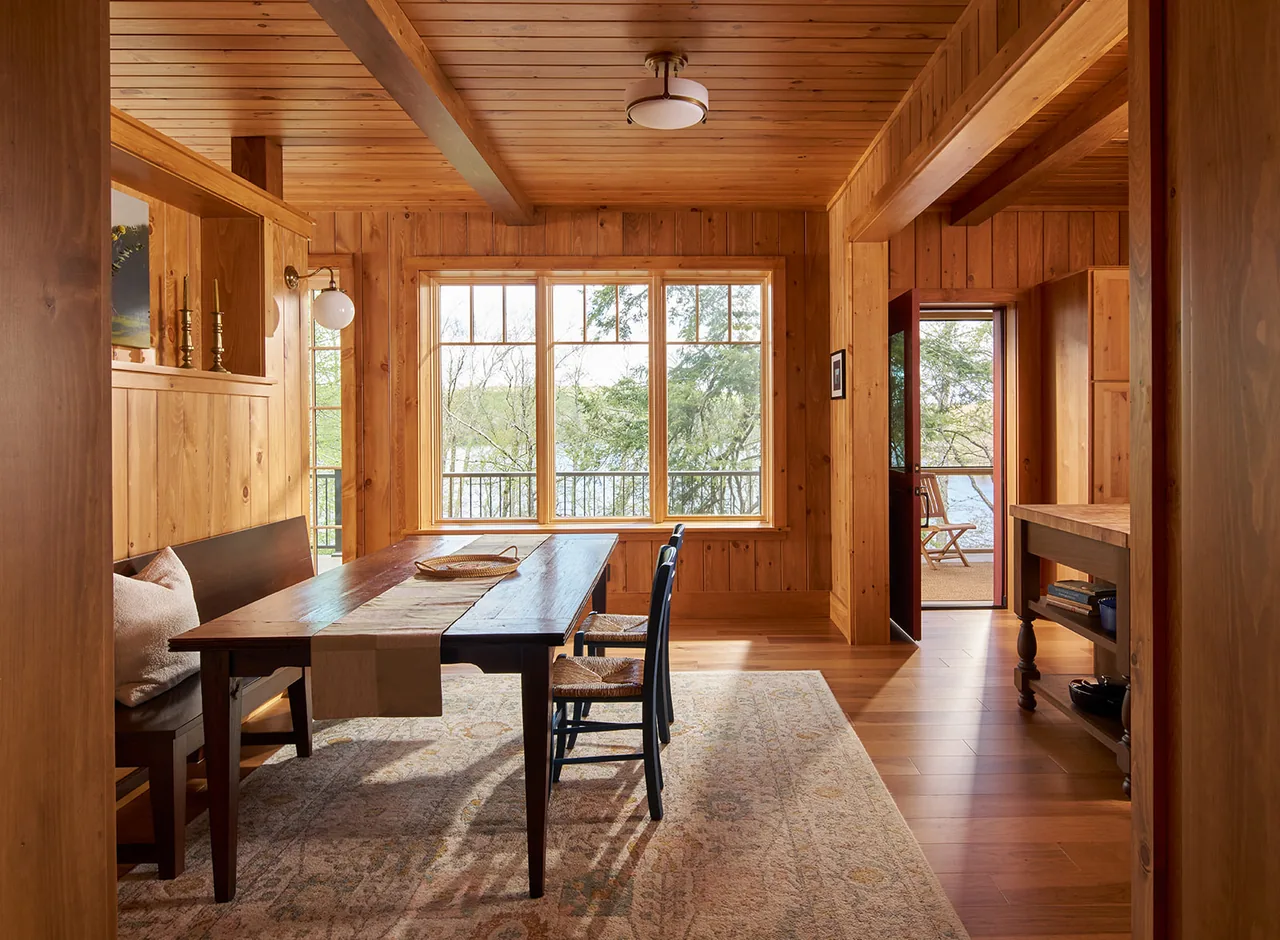 The Lake Cottage dining area features wood paneling, a window with a view of surrounding trees, and simple wooden furniture.
