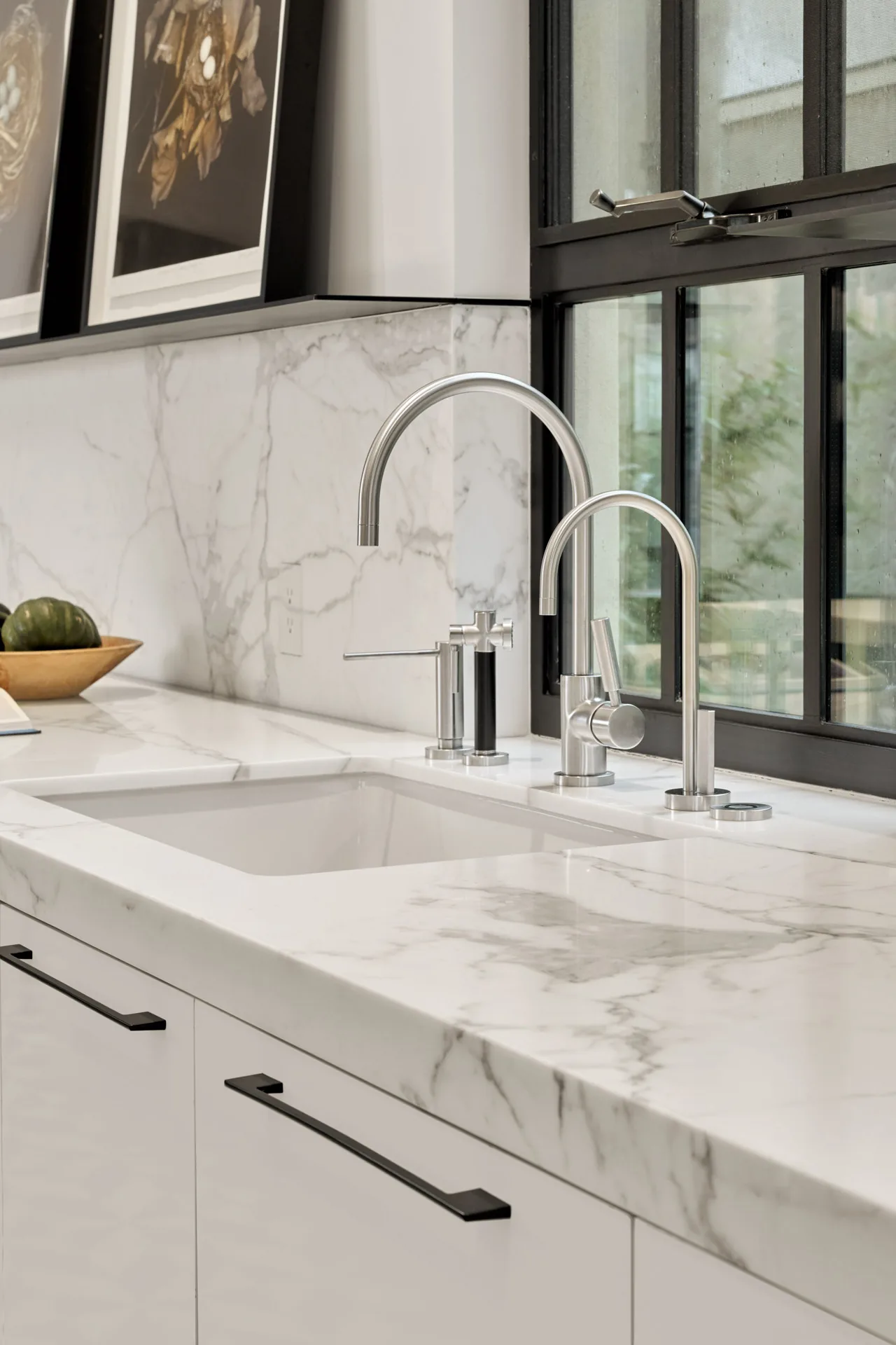 Kitchen sink area featuring white marble surfaces, dual stainless steel faucets, and a black-framed window.