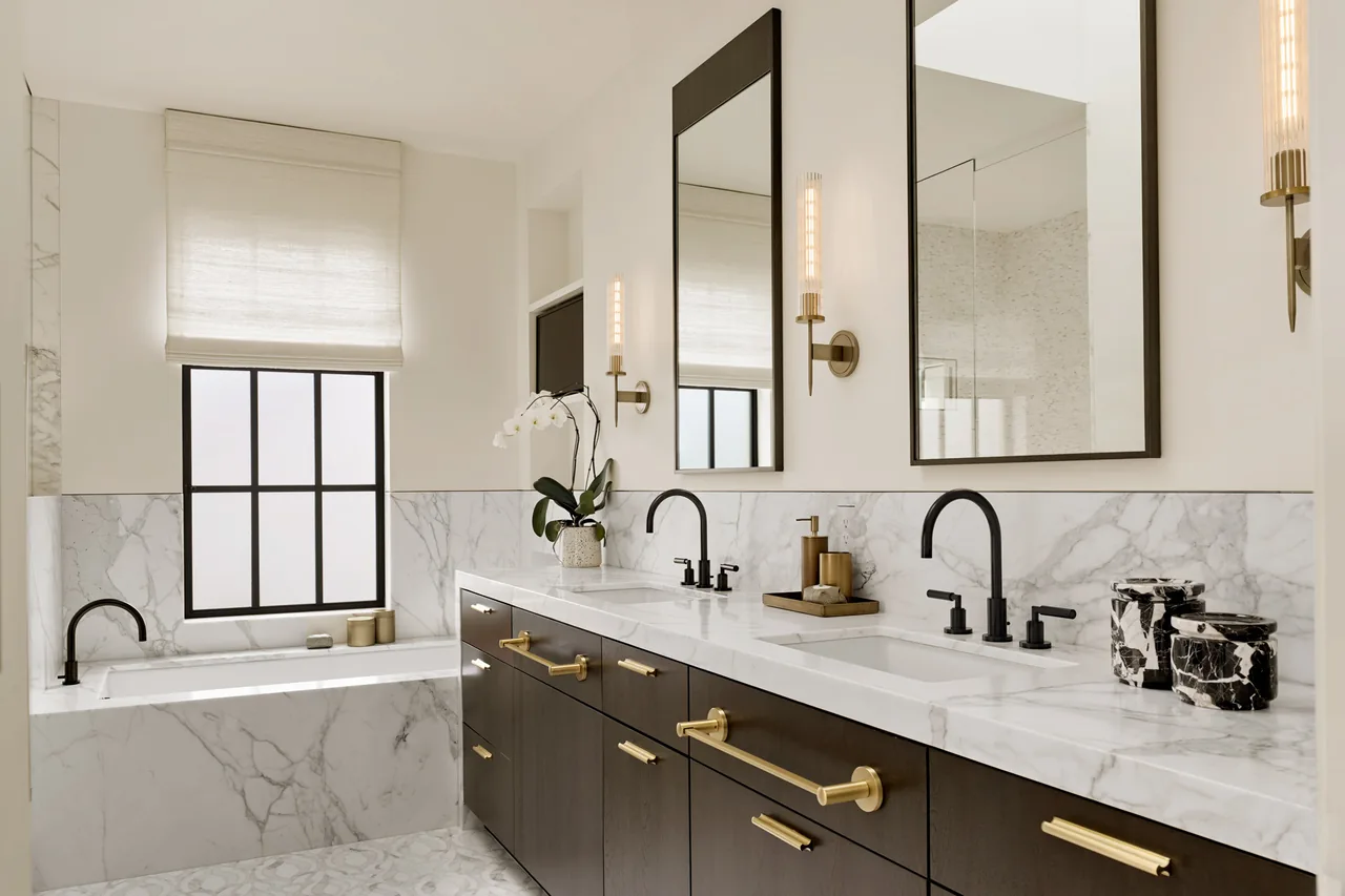 Bathroom features dual black faucets, marble backsplash, and dark cabinets with gold pulls and hardware.