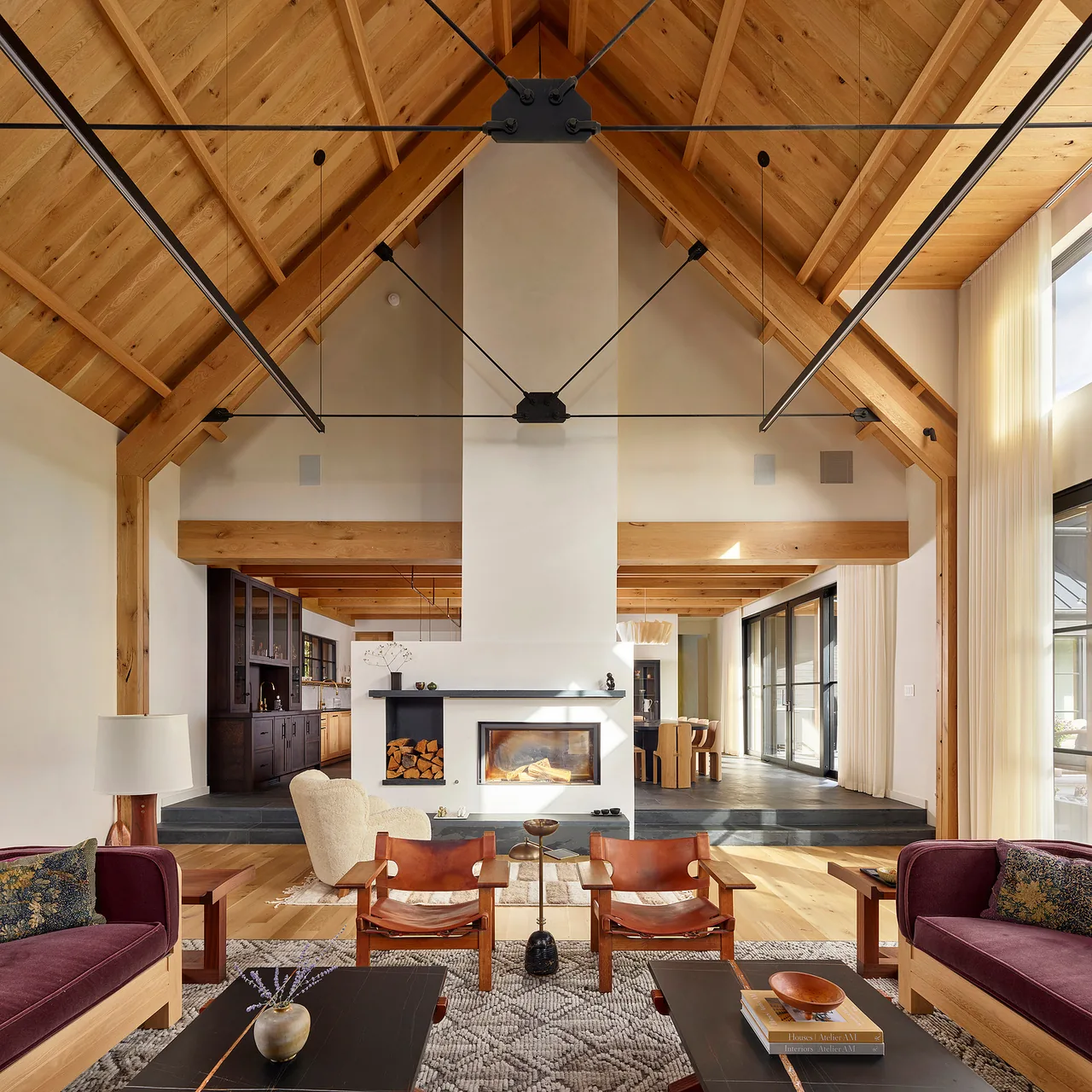 Living room with exposed wood ceiling beams, a fireplace centered on a white wall, and large windows with light curtains.