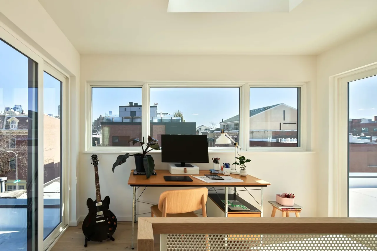 Home office in the Two Door House features a wooden desk and chair positioned in front of three windows with urban views.