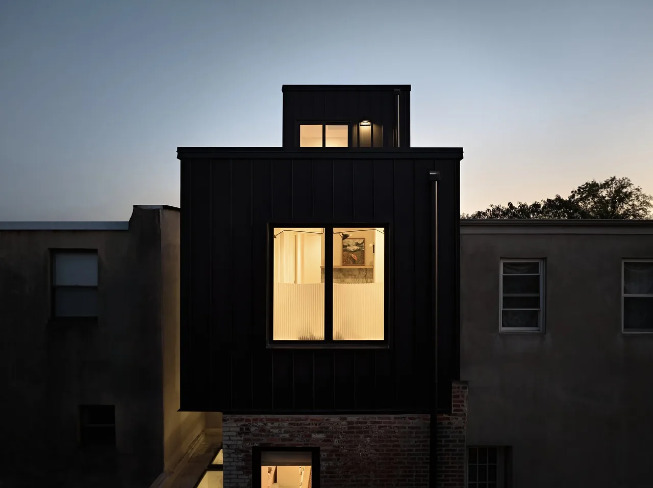 Black panel exterior with large window shows two-story extension on older brick structure at dusk.