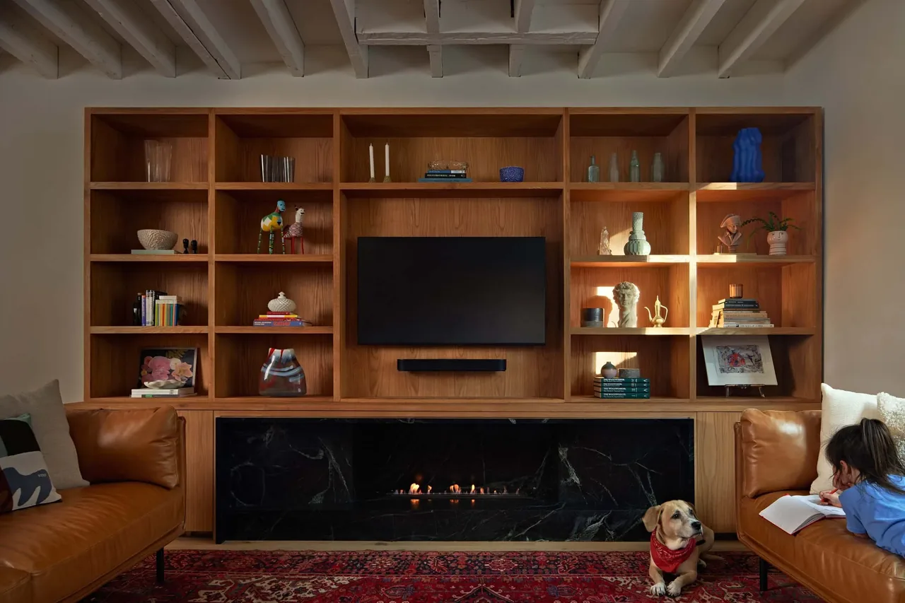 Living room featuring a built-in wooden shelving unit around a mounted TV and a black linear fireplace below, with exposed ceiling beams.