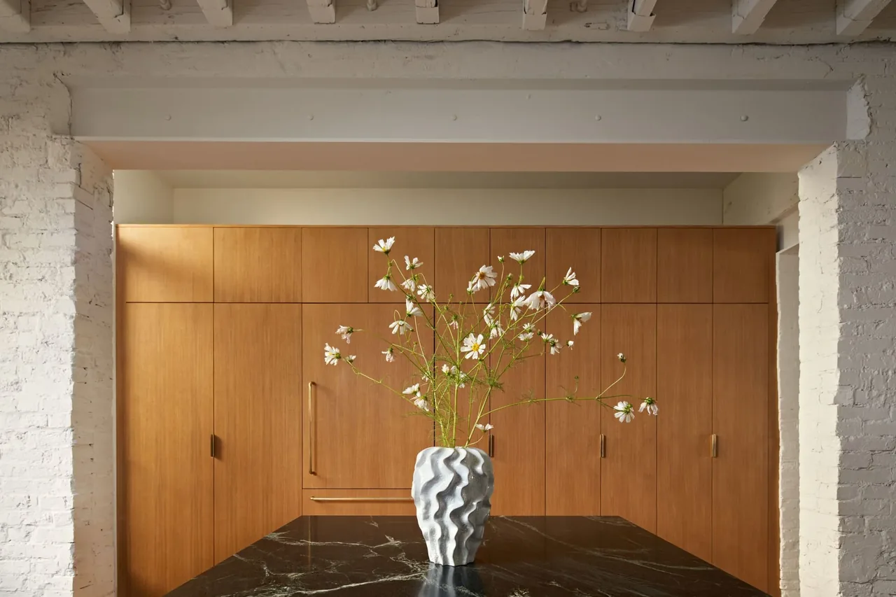 Wood-paneled cabinetry with wood handles fronts a kitchen area opposite a black marble countertop with a textured flower vase.