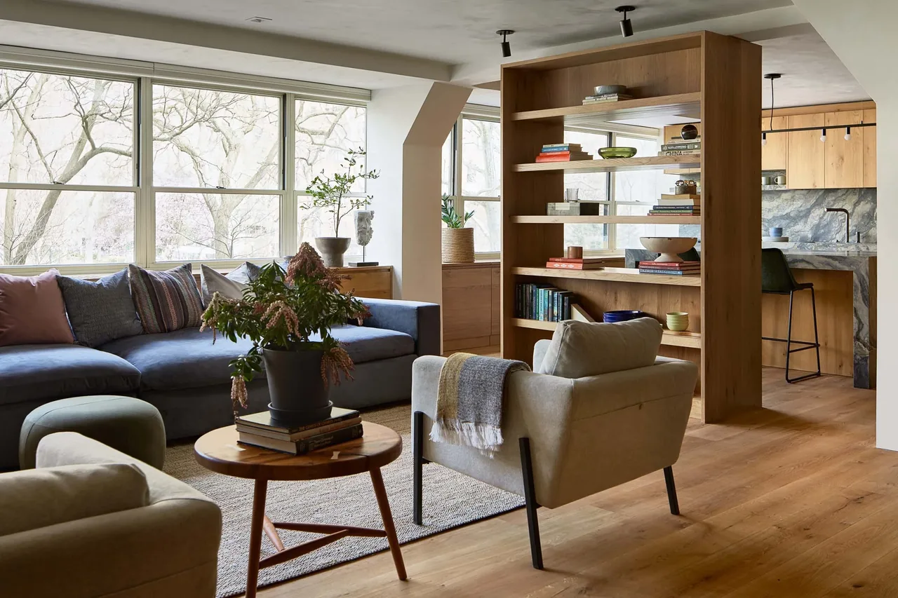 Open living room with blue sofa and wood shelving unit dividing space from kitchen with wood cabinetry and stone backsplash.