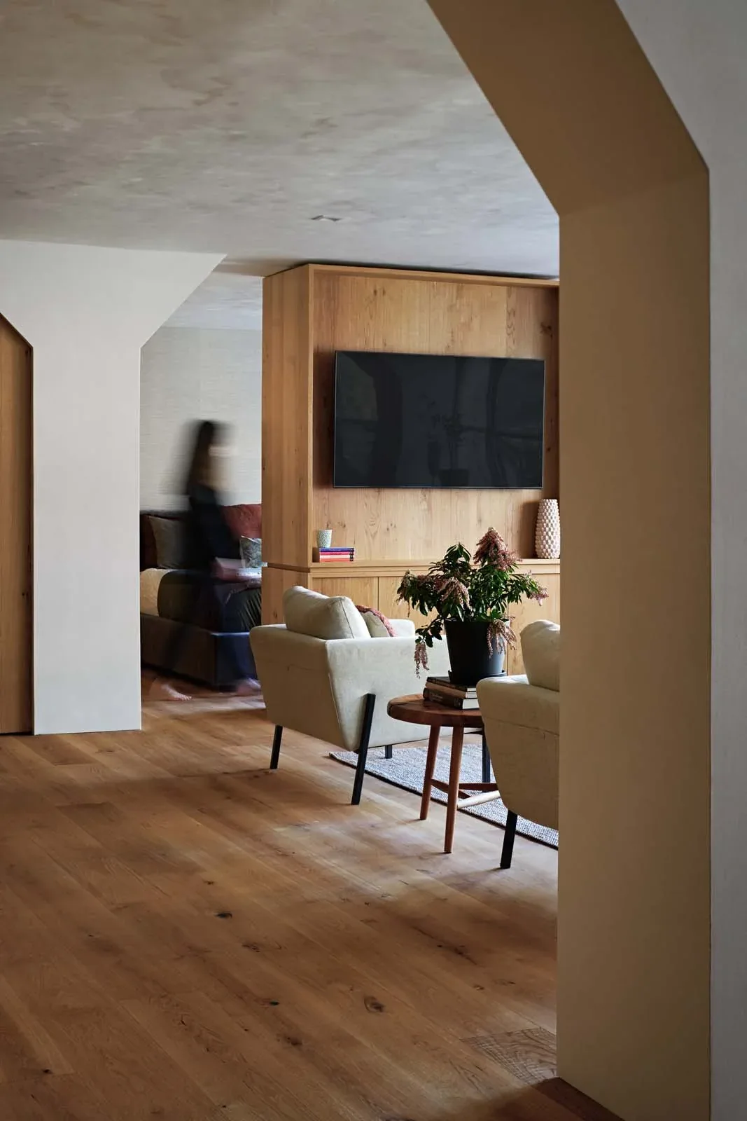 The Low Penthouse features a warm living space with a wood-paneled media wall and light upholstered armchairs.