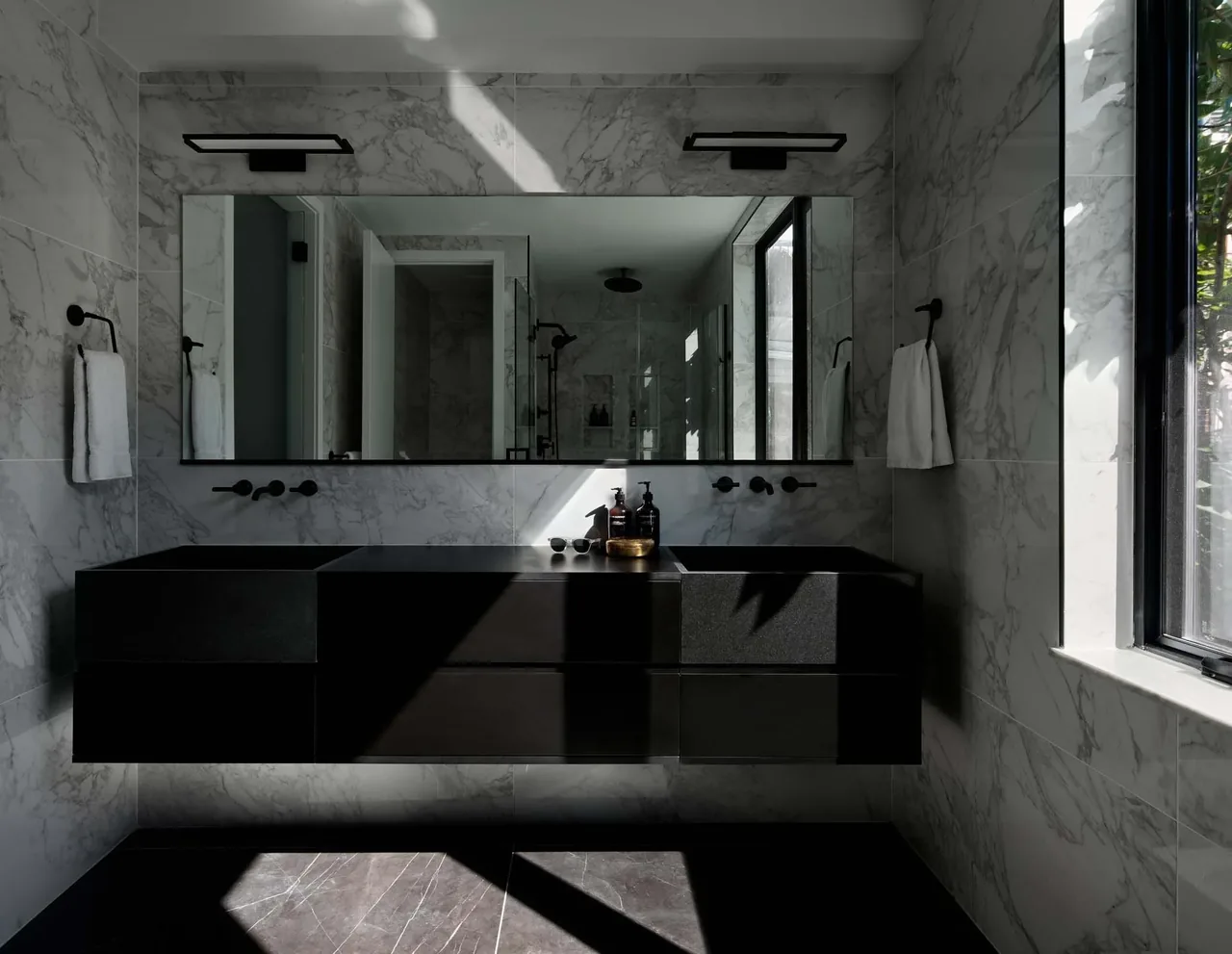 Dual black sinks with wall-mounted black fixtures under a large horizontal mirror in a marble-clad bathroom.