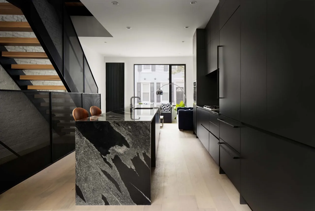 The Contextual House kitchen features black flat-panel cabinets, a large marble island, and wooden bar stools alongside a black metal staircase.