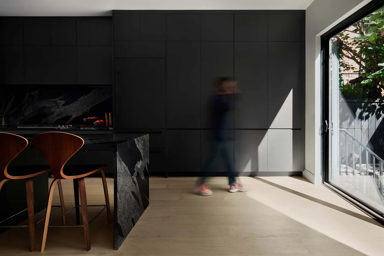 The Contextual House kitchen features dark cabinetry, a stone island, and wood bar stools with natural light from sliding glass doors.