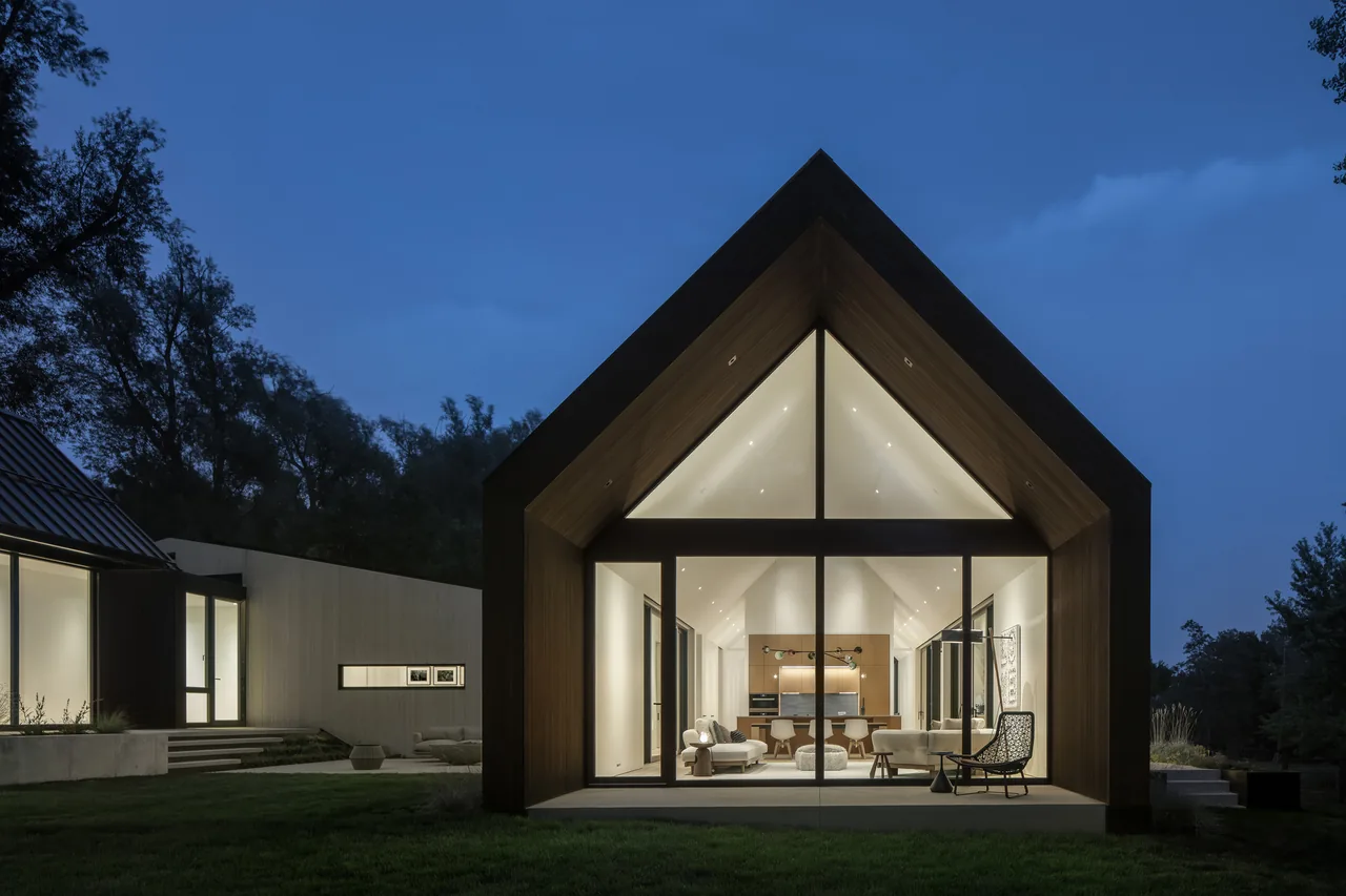 Three Gables project features a modern house with a prominent gable and large glass windows illuminating the interior living area at night.