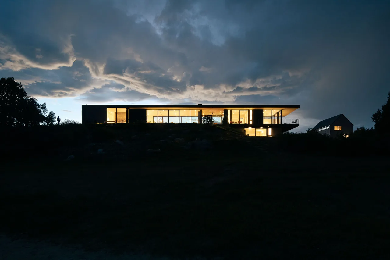 Rivers Edge house by Piché Architecture features illuminated floor-to-ceiling glazing in a modern design at dusk.