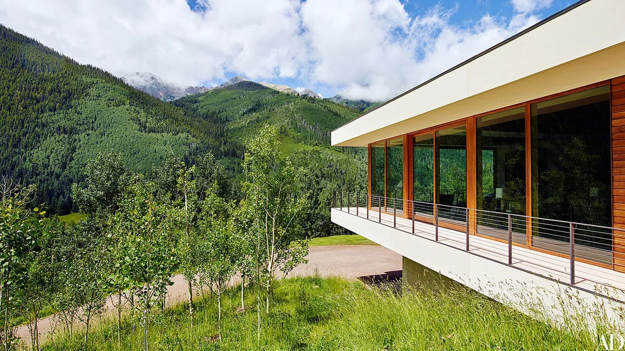 Modern house with floor-to-ceiling wood-framed windows and a cantilevered balcony overlooking a lush green mountainous landscape.