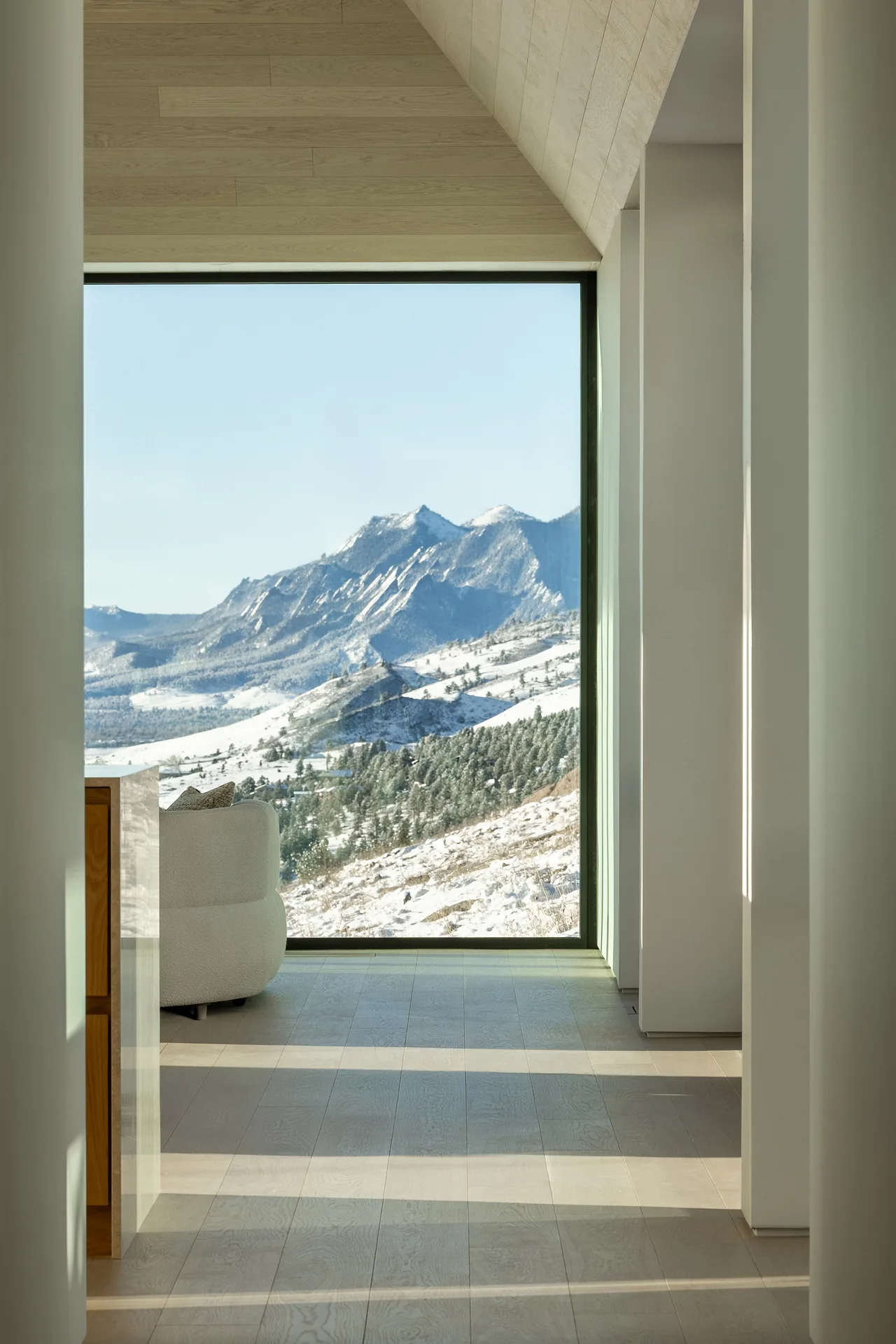 Interior view shows a large picture window framing snowy mountains beyond a light wood floor and walls.
