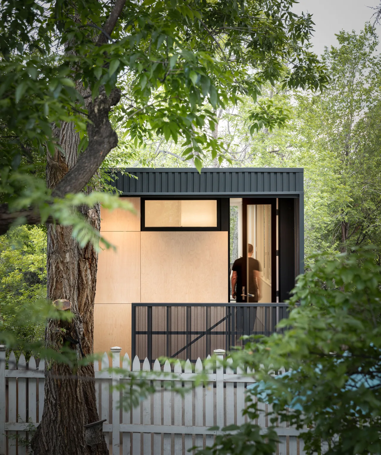 The Space Pod project by Piché Architecture features vertical metal cladding and wood paneling, visible behind a white picket fence and trees.