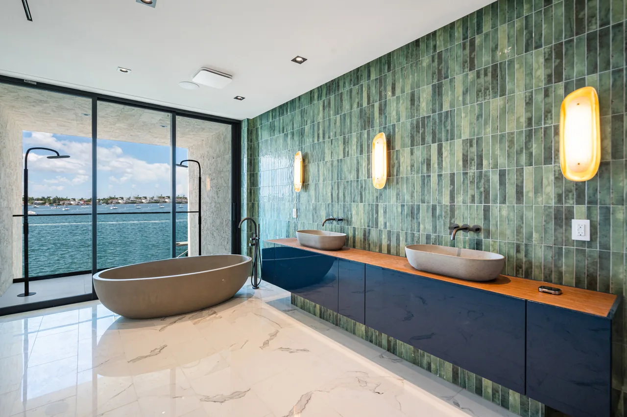 The Venetian Island II bathroom features green tiles, a freestanding tub, double vessel sinks, and a waterfront view through floor-to-ceiling glass.