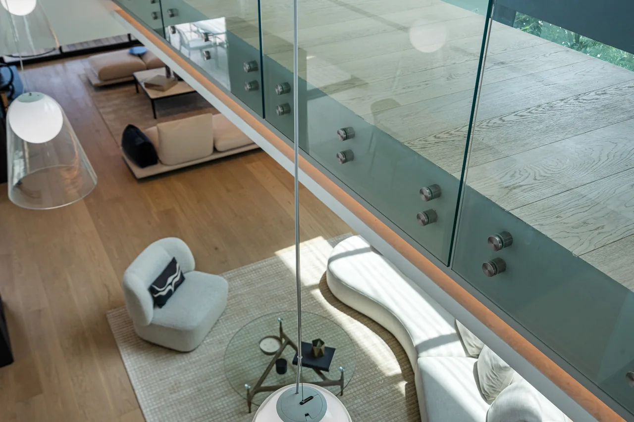 A modern living room with glass balcony railing, light wood floors, and curved sectional sofa under soft natural light.