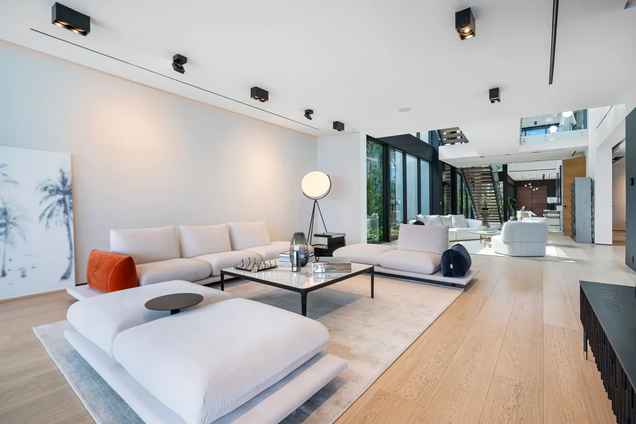 Open living area featuring low-profile white sofas, light wood floors, and black frame floor-to-ceiling windows by a modern staircase.