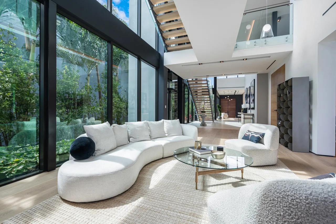 Living room with curved modular sofa, glass coffee table, and large black-framed windows revealing lush outdoor greenery.