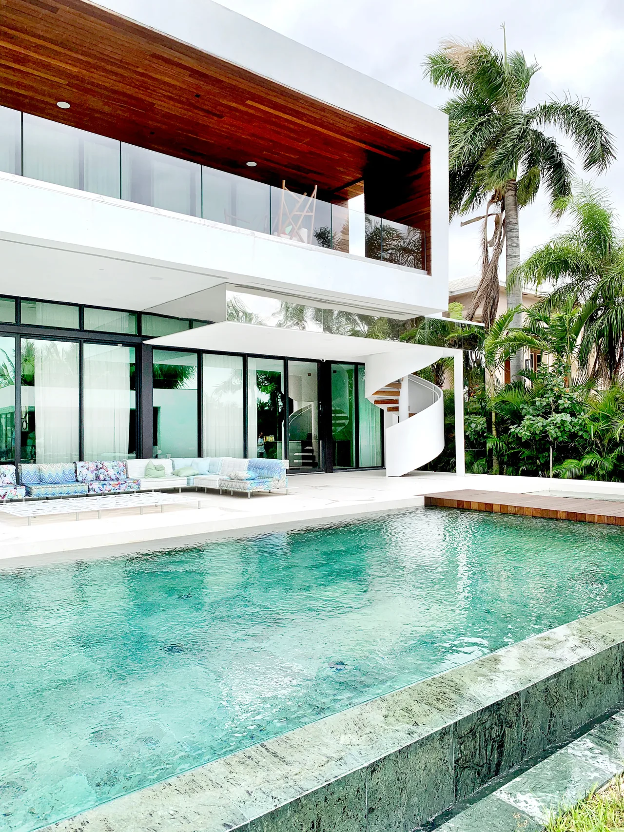 Exterior of Palm Island Miami home features glass walls, a wood-ceiling balcony, and a pool with a spiral staircase.