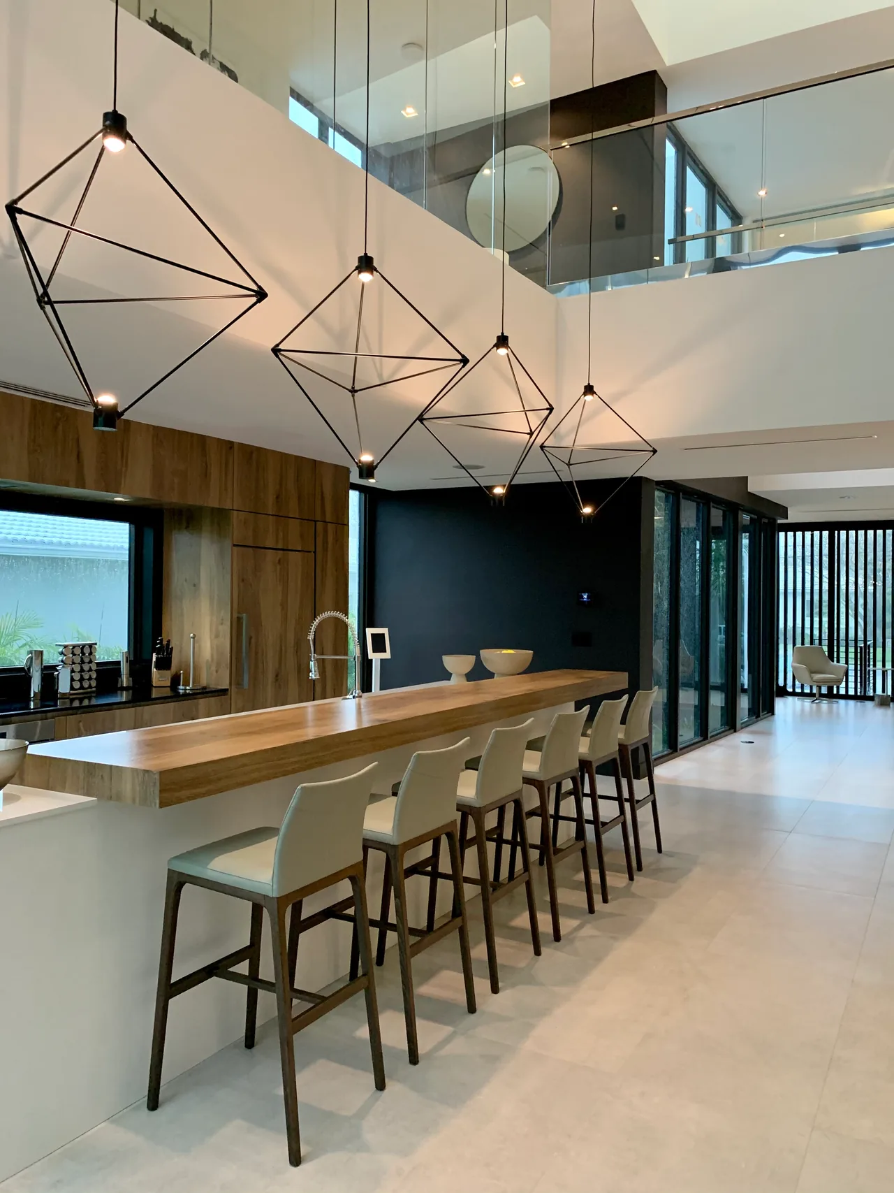 Open kitchen bar with wood countertop, geometric pendant lights, and floor-to-ceiling glass in a modern style home.