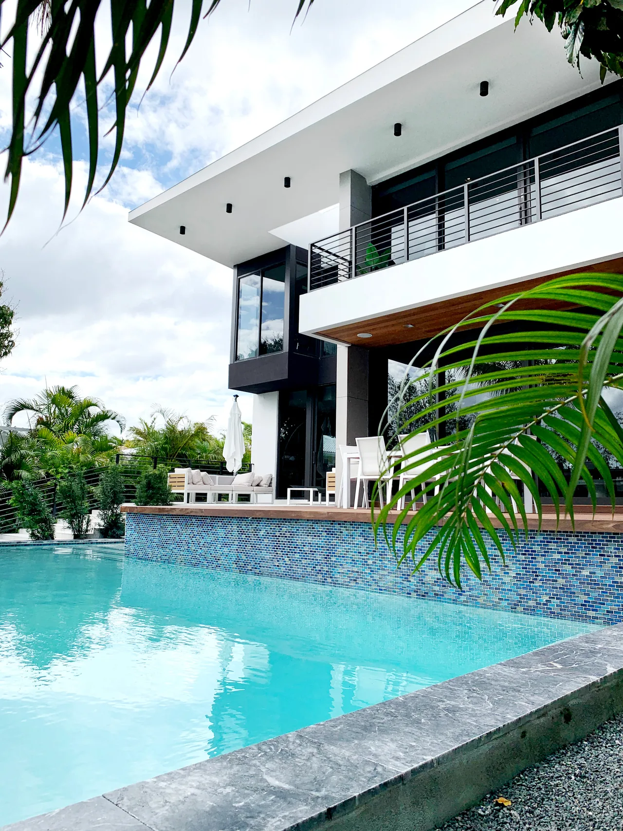 Modern two-story home exterior with balcony overlooking a pool and patio seating area.