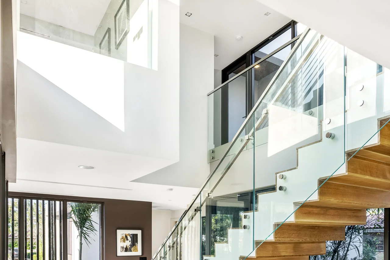 Modern staircase with wooden steps and glass railings under a white ceiling featuring a large square cutout.