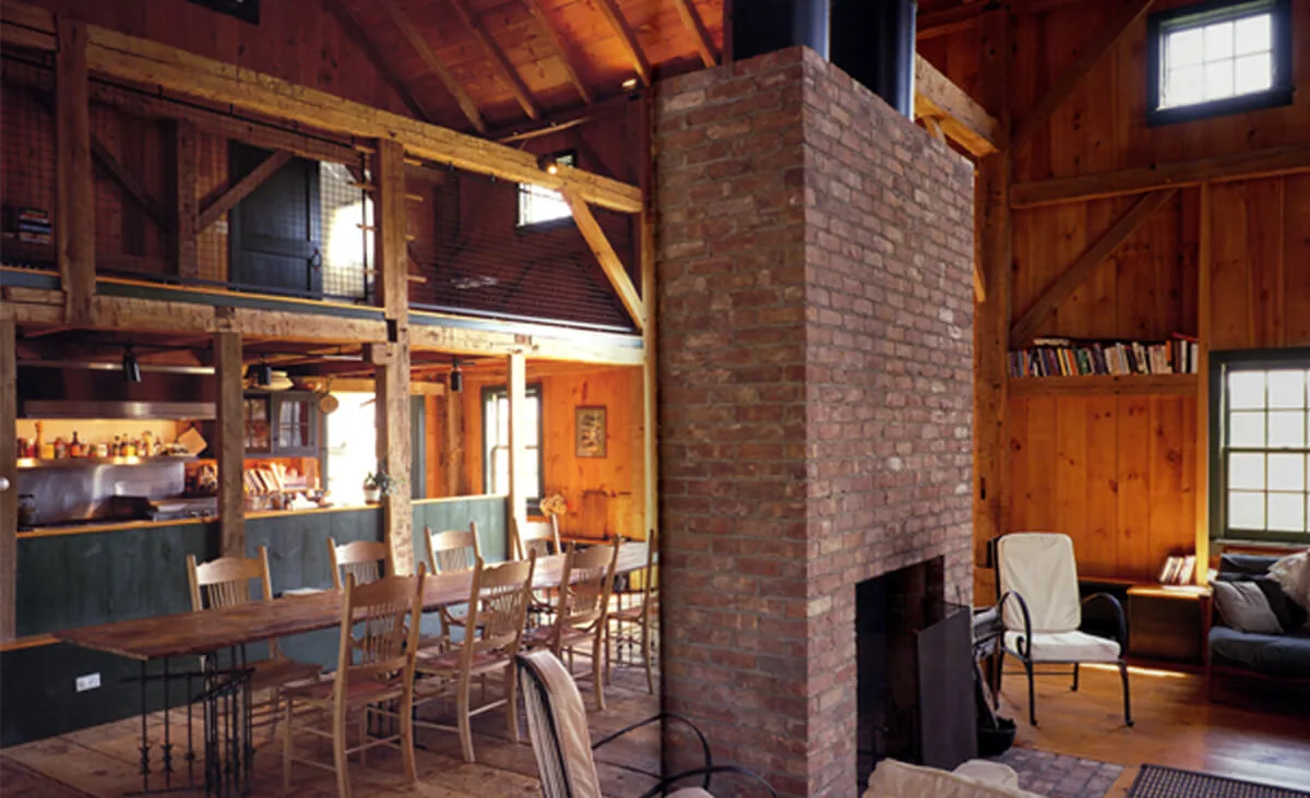 Middleton-Waln Barn Home interior shows an open dining and living space with exposed wooden beams and a central brick fireplace.