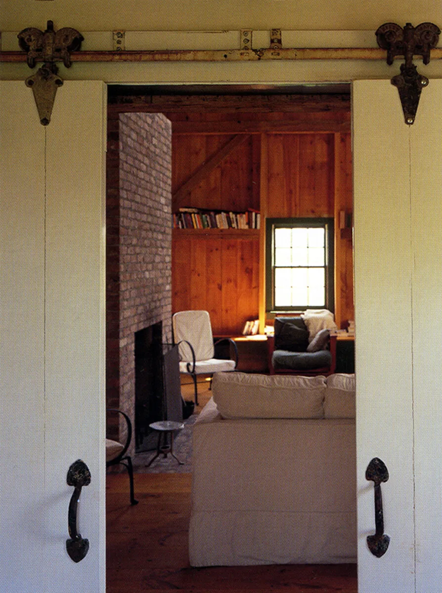 Interior view through barn-style sliding doors into a living room with wood paneling and a brick fireplace in the Middleton-Waln Barn Home.