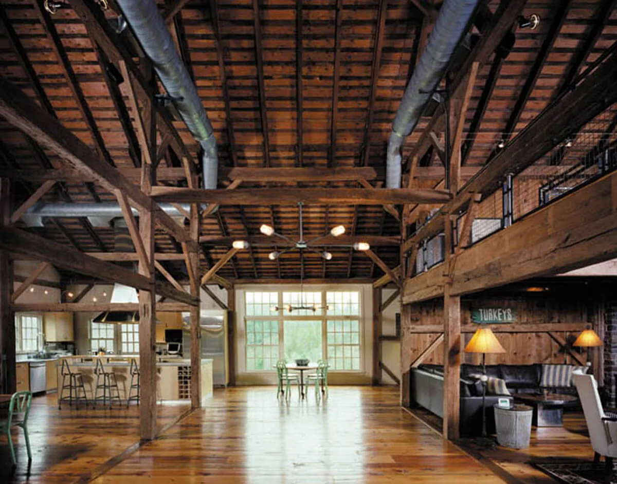 Open barn home interior featuring exposed wood beams, a rustic dining table, and large multi-pane windows.