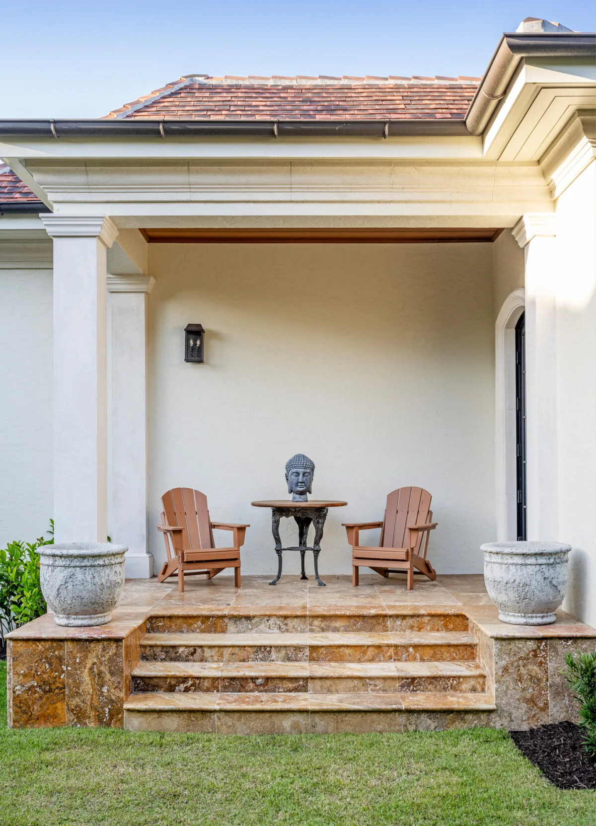 A covered patio with stone steps features wooden chairs, a small table with a Buddha head sculpture, and a lantern-style wall sconce on the wall.