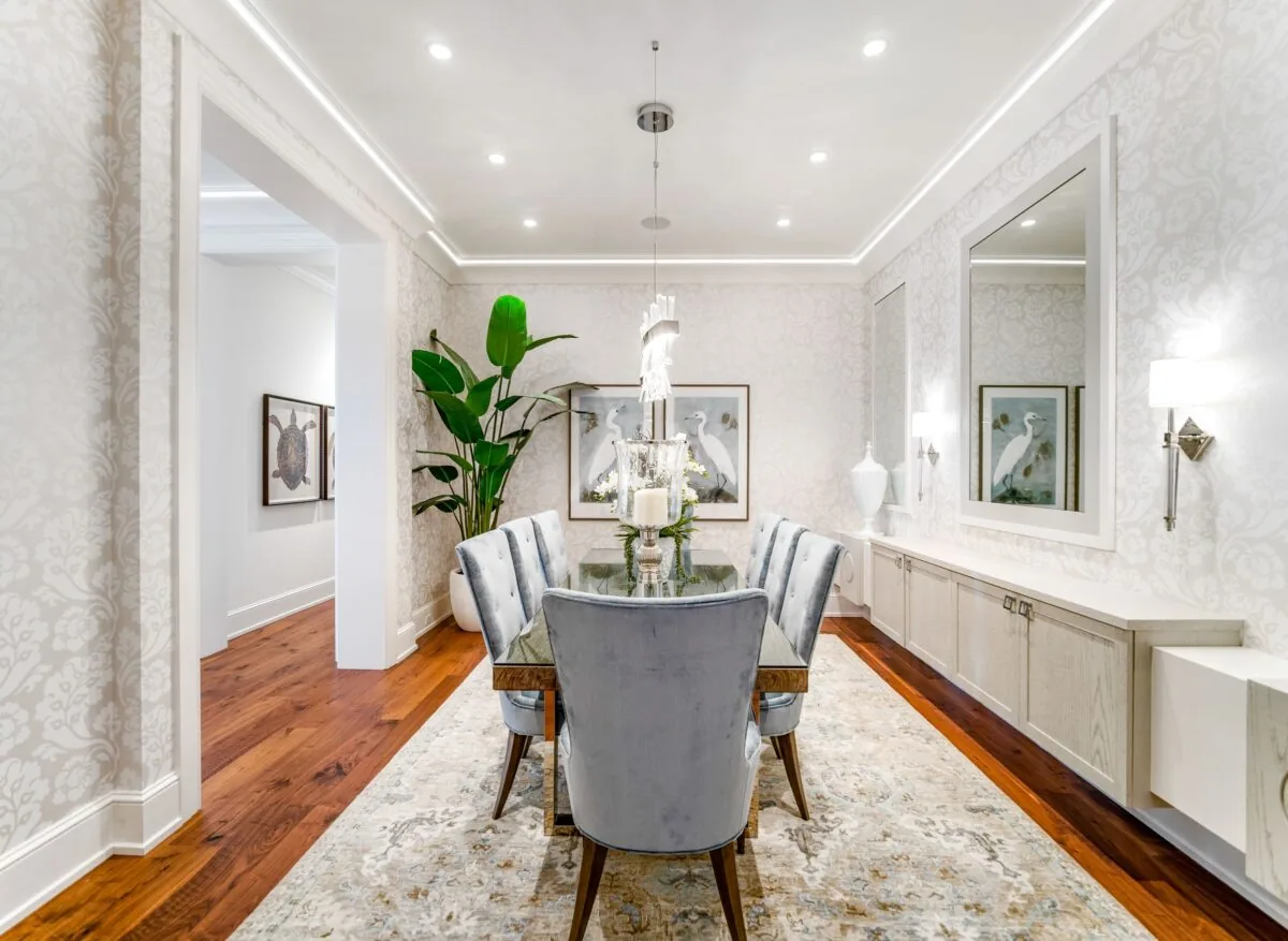Dining room featuring a glass table with six velvet chairs and floral wallpaper, accented by a large indoor plant and wall art.