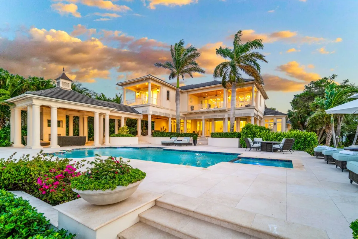 Two-story home with a pool and outdoor seating next to lush landscaping and palm trees.