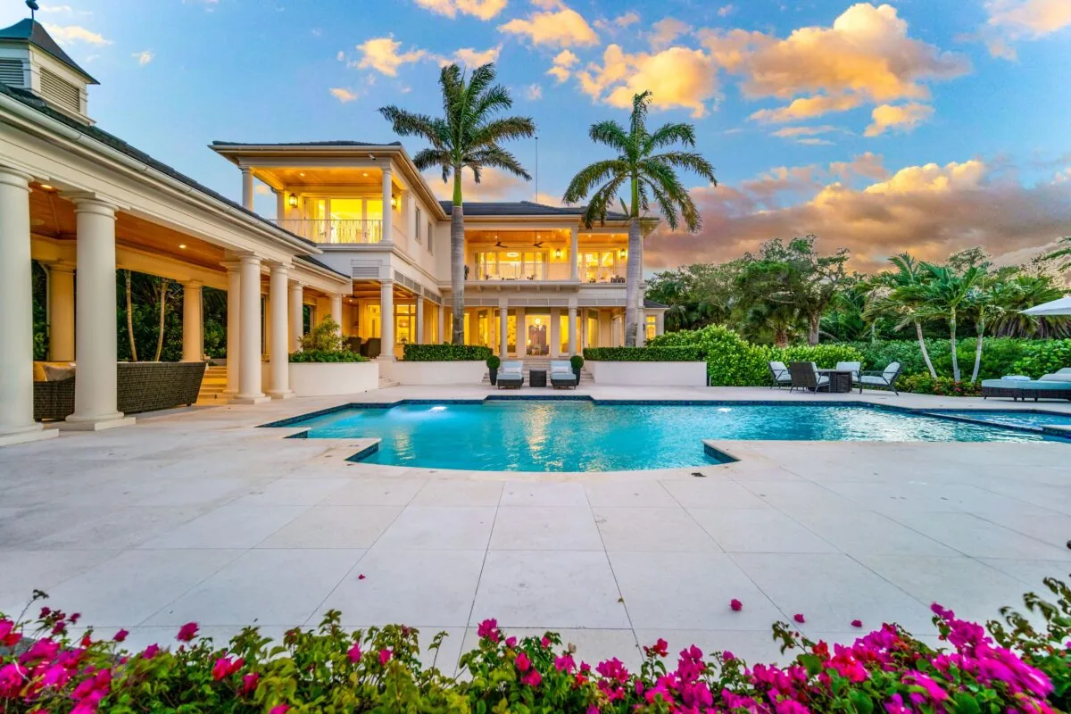 Bayfront residence with a large pool, surrounded by lush landscaping and two tall palm trees at sunset.