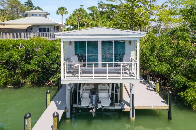 The Tide House boathouse features a covered balcony with seating and a boat lift below, adjacent to a dock over the water.