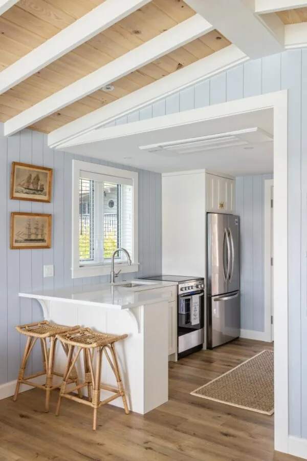 The kitchen of Tide House features light blue walls, white cabinetry, wood floors, and a vaulted ceiling with exposed beams.